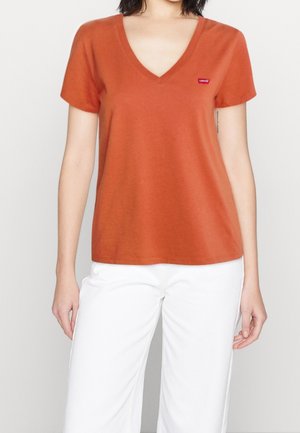 Woman wearing a rust-orange V-neck short-sleeve T-shirt and white pants, standing against a plain light background.