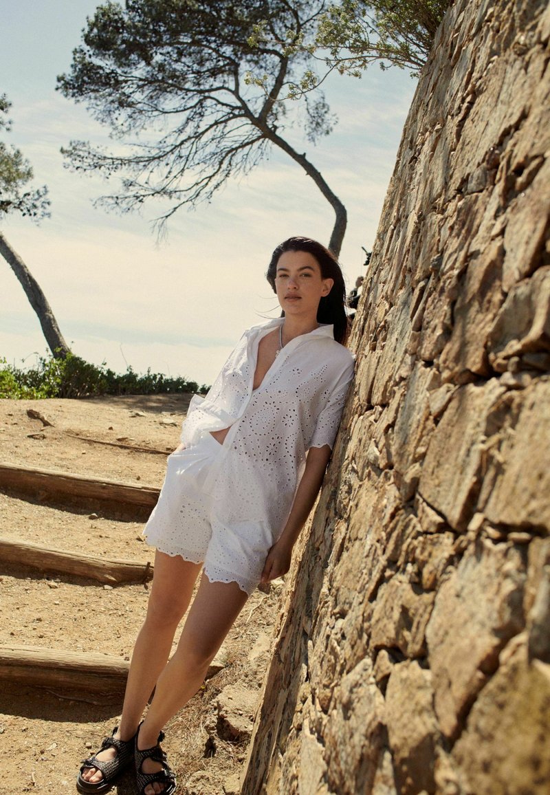 Set di camicia e pantaloni corti in broderie anglaise bianca con bordi scallop, abbinato a sandali neri testurizzati su uno sfondo di parete rocciosa.