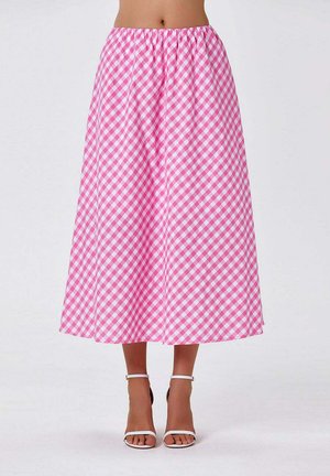 Femme portant une jupe midi à carreaux roses et blancs et des talons blancs ouverts avec bride à la cheville, debout devant un fond uni.