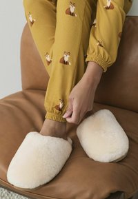 Baltos pūkuotos šlepetės su suapvalinta forma ant rudas odinės kėdės; asmuo reguliuoja pižamos rankoves su gelsvais lapės raštais.