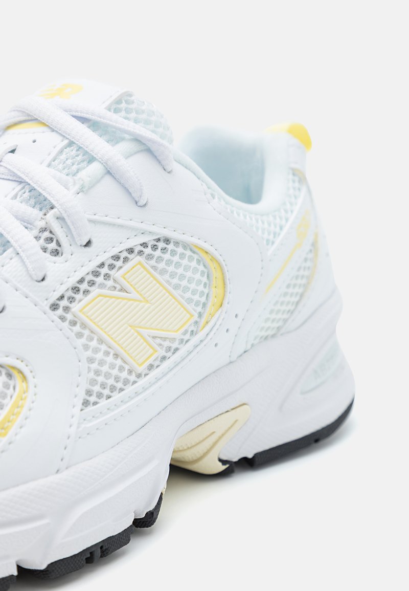 Białe siatkowe sneakersy z wyraźnym żółtym logo, sznurowanym zapięciem, teksturowanym noskiem oraz wyściełaną podeszwą z czarnymi akcentami.