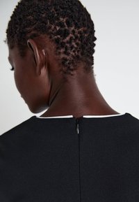 Μαύρο φόρεμα με λευκό λαιμό και κεντρικό φερμουάρ στο πίσω μέρος. Το ύφασμα φαίνεται ομαλό με ματ φινίρισμα. Κοντινή λήψη από πίσω.