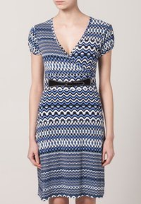 Niebieska i biała sukienka w wzór zygzakowy z dekoltem w serek, krótkimi rękawami i czarnym paskiem podkreślającym talię. Wykonana z elastycznego materiału.