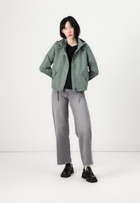 Green, lightweight jacket with a hood, snap buttons, and side pockets, paired with gray wide-leg jeans and black shoes.