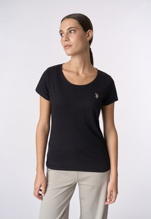 Donna con i capelli castani raccolti in una coda di cavallo, che indossa una maglietta nera a maniche corte con un piccolo logo ricamato e pantaloni beige, che guarda a sinistra.