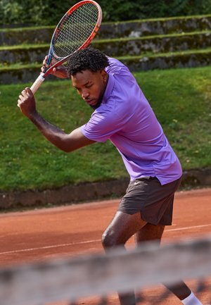 Tennis spiller i en lilla kortærmet skjorte og brune shorts, som holder en rød og blå racket og forbereder sig på at ramme en tennisbold på en grusbane.