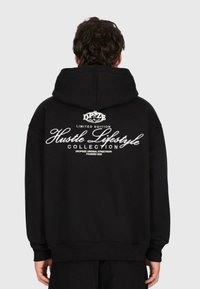 Eine Person trägt einen schwarzen Kapuzenpullover mit weißem Text "Hustle Lifestyle Collection, Limited Edition" auf dem Rücken und steht vor einem schlichten Hintergrund.