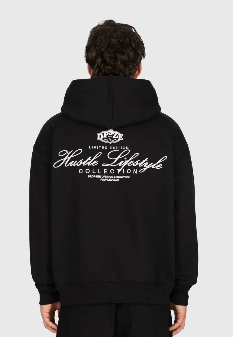Eine Person trägt einen schwarzen Kapuzenpullover mit weißem Text "Hustle Lifestyle Collection, Limited Edition" auf dem Rücken und steht vor einem schlichten Hintergrund.