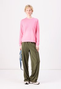 Roze sweatshirt met een ronde halslijn, gecombineerd met olijfgroene, losse broeken. Model houdt een blauwe schoudertas vast; draagt lichte sneakers.