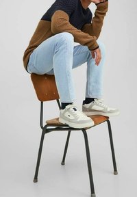 Zapatillas para hombre en gris claro con acentos en blanco; parte superior texturizada, lengüeta acolchada y cierre de cordones. Combinadas con jeans de color azul claro y un suéter de punto marrón.