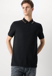 Cămașă polo neagră cu mâneci scurte, având un guler maro în contrast, placket cu trei butoane și brand subtil pe piept.