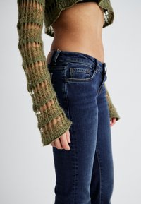 Olivenfarget strikket crop top med åpen vevdesign og bell sleeves, parret med mørkeblå tettsittende jeans med subtile sømdetaljer.