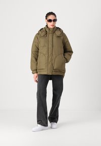 Casaco acolchoado verde-oliva com colarinho alto e capuz, apresentando grandes bolsos frontais, usado sobre uns jeans pretos de corte largo e ténis brancos.