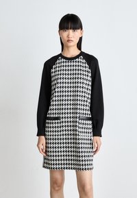 Mustavalkoinen houndstooth-mekko, jossa on mustat pitkät hihat, kaksi etutaskua ja pyöreä pääntie. Vaatteessa on sileä rakenne ja rento muotoilu.