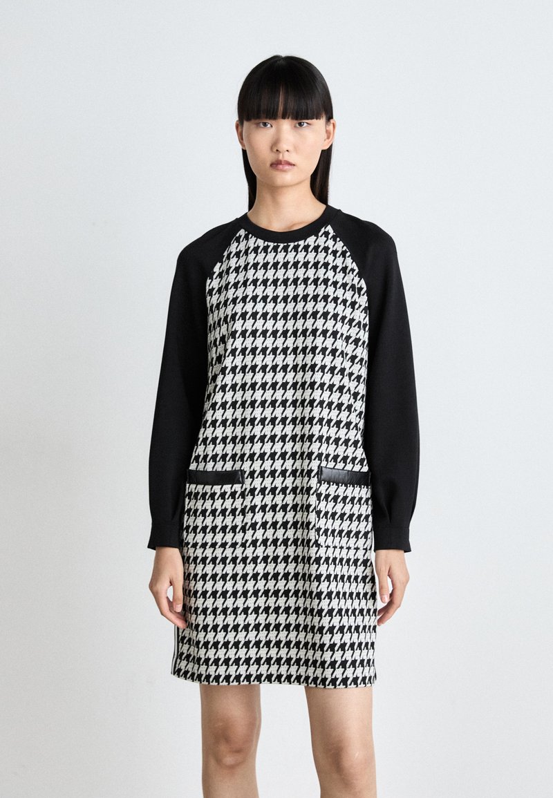 Mustavalkoinen houndstooth-mekko, jossa on mustat pitkät hihat, kaksi etutaskua ja pyöreä pääntie. Vaatteessa on sileä rakenne ja rento muotoilu.
