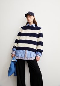 Pull en tricot rayé bleu marine et blanc superposé sur une chemise à rayures bleu clair, associé à un pantalon large noir et une casquette bleu marine.