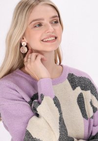 Pulover violet și crem cu accente negre, având o croială relaxată și manșete ribbate. Modelul poartă cercei circulari mari și un inel.