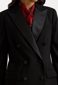 Melna divrindas žakete ar samta apmalēm, lieliem apaļiem pogām un sarkanas uzraksta šalli, kas sasieta ap apkakli. Tekstūrēta auduma akcenti.