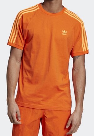 Osoba ubrana w jaskrawo pomarańczową koszulkę Adidas z trzema jaśniejszymi pomarańczowymi paskami na rękawach i małym logo Adidas na lewej piersi.