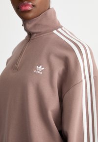 Sweatshirt castanho com fecho éclair e gola alta, apresentando o logótipo branco da Adidas e três riscas brancas ao longo dos ombros. Textura de tecido suave.