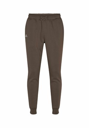 Homme portant un pantalon de jogging marron avec cordon de serrage et des baskets blanches, debout devant un fond uni.