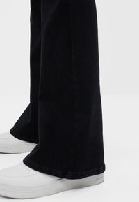 Schwarze ausgestellte Jeans mit einem sauberen Saum, kombiniert mit weißen Sneakers. Der Stoff wirkt robust und hat eine glatte Textur.