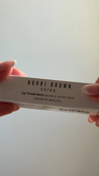 Des mains aux ongles longs tiennent une boîte blanche étiquetée Bobbi Brown Extra Lip Tinted Balm avec un poids produit de 0,08 oz/2,5 g.