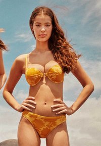 Žluté bikiny s květinovým vzorem, které mají vycpanou podprsenku s centrální mašlí a odpovídající nízké kalhotky. Hladký textura látky.