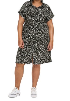 Kobieta w czarnej sukience w białe abstrakcyjne kropki, z krótkimi rękawami, zapinanej na guziki z przodu, w białych trampkach, stojąca na białym tle.