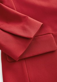Rød tekstil med en struktureret overflade, der har en struktureret ærme og en indsætslomme. Stoffet virker holdbart og glat.