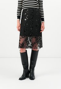 Kvinne iført svart blonde skjørt med blomsterpailleter, svarte høye støvler, og en langermet topp med svart og hvit striper.