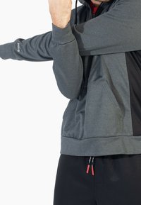 Felpa atletica grigia con dettagli neri, texture morbida, tasche con zip e cordoncino. Presenta un logo sulla manica.