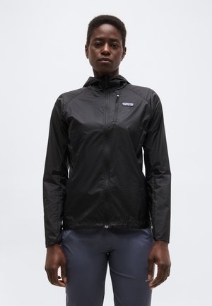 Άτομο που φοράει μαύρο φούτερ με φερμουάρ και κουκούλα και σκούρο παντελόνι, στέκεται μπροστά σε μια απλή ανοιχτό γκρι φόντο.