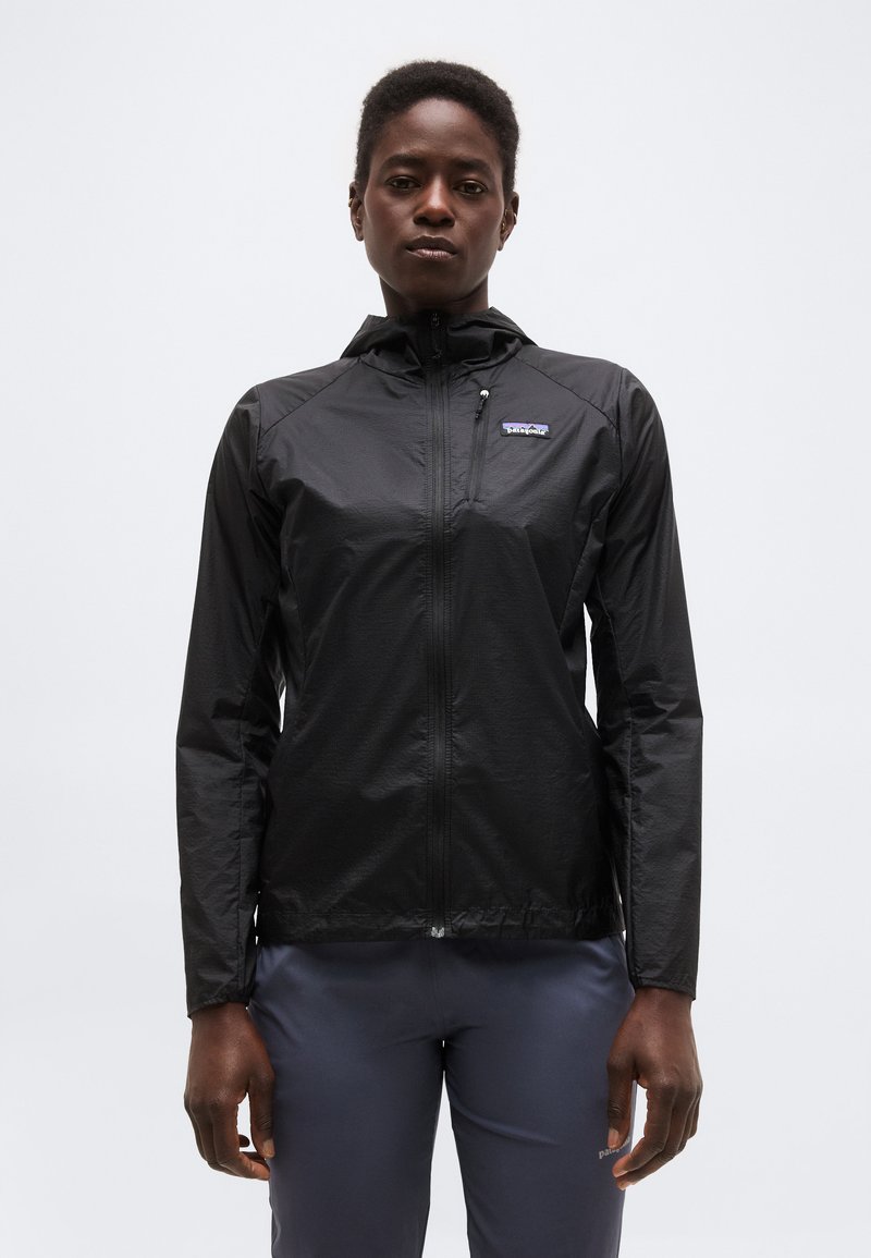 Person iført en sort hættejakke med lynlås og mørke bukser, der står foran en ensartet lysegrå baggrund.