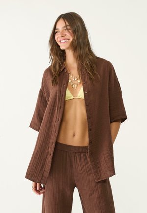 Braune Button-Down-Bluse aus leichtem strukturiertem Stoff, mit kurzen Ärmeln und lockerer Passform, kombiniert mit passenden braunen Hosen.