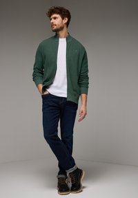 Cardigan verde strutturato sopra una maglietta bianca, abbinato a jeans blu scuro e stivaletti neri con suola beige, posato su uno sfondo neutro.