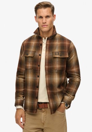 Chemise en flanelle à carreaux marron avec des boutons noirs, deux poches sur la poitrine, col et manches longues, portée par-dessus un haut crème.
