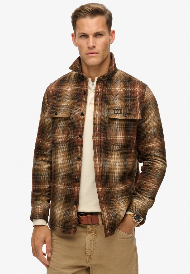 Brunternet flannel skjorte med sorte knapper, to brystlommer, krave og lange ærmer, båret over en cremefarvet top.