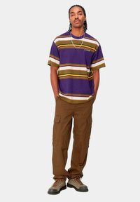 Camiseta morada a rayas con tiras verdes, amarillas y blancas, combinada con pantalones cargo marrones y botas de senderismo marrones.