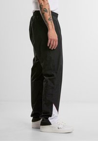 Pantalons de training en nylon noirs avec une texture lisse, présentant une bande d'accent blanche et rouge au bas. Taille élastique et coupe décontractée.