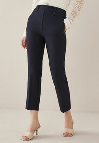 Donna che indossa pantaloni neri aderenti a lunghezza caviglia, blouse bianca in pizzo a maniche lunghe e sandali con tacco aperto beige, in piedi contro uno sfondo neutro.