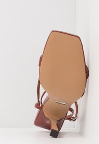 Sandale à talon haut en cuir brun vue de la semelle, montrant un bout carré, un talon fin et une bride cheville avec boucle.