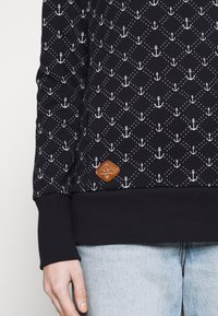 Gros plan d'une chemise à manches longues foncée avec un motif d'ancres blanches et de lignes pointillées, portée avec un jean bleu clair.