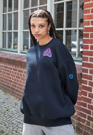 Sort oversized sweatshirt med rund halsudskæring og sænkede skuldre. Har et flerfarvet geometrisk grafisk motiv på brystet og et blåt accentlogo.
