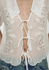 Blouse en lin blanc avec broderie florale, décolleté en V profond avec liens et ourlet festonné. Assortie avec un short en jean.