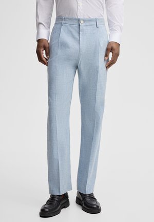 Man draagt lichtblauwe broek met plooi, zwarte loafers en een wit overhemd met lange mouwen, staand tegen een effen achtergrond.