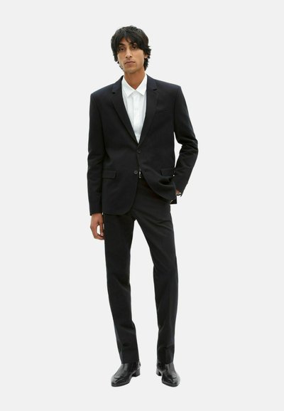 Schwarzer maßgeschneiderter Anzug mit einreihigem Blazer und glatten Hosen. Kombiniert mit einem weißen Hemd mit Kragen und glänzenden schwarzen Schuhen.