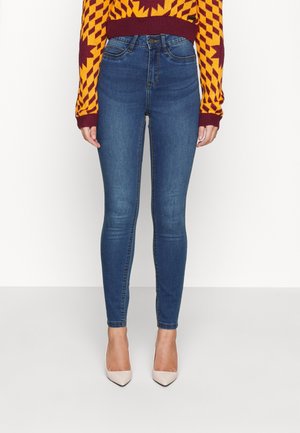 Femme portant un jean skinny bleu taille haute, un pull orange et bordeaux à motifs et des talons pointus beige, debout devant un fond blanc.