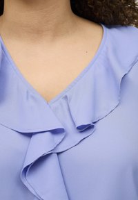 Helllila Bluse mit V-Ausschnitt, zarten Rüschendetails am Kragen und glatter, fließender Stofftextur.