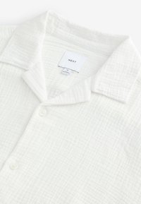 Camicia bianca button-up con un tessuto texturizzato e stropicciato. Presenta un colletto classico e una chiusura con un solo bottone, etichetta visibile all'interno.
